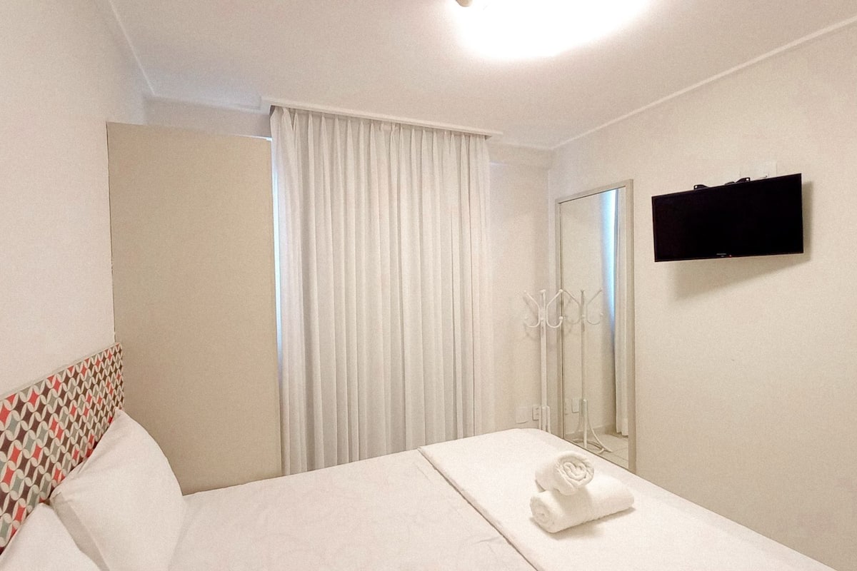 Quarto de hotel com cama, televisão, espelho e janela com cortinas.