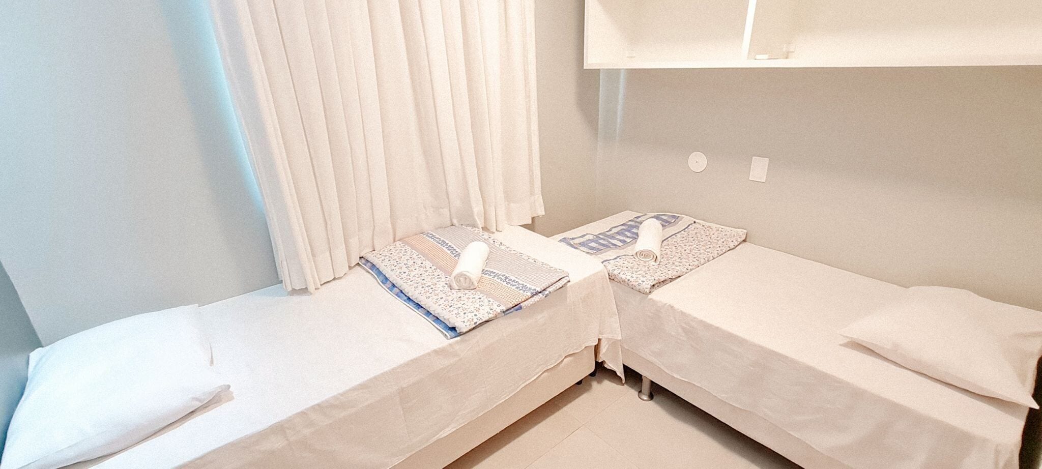Two single beds with white linens, a window with white curtains, and a wall-mounted light fixture.