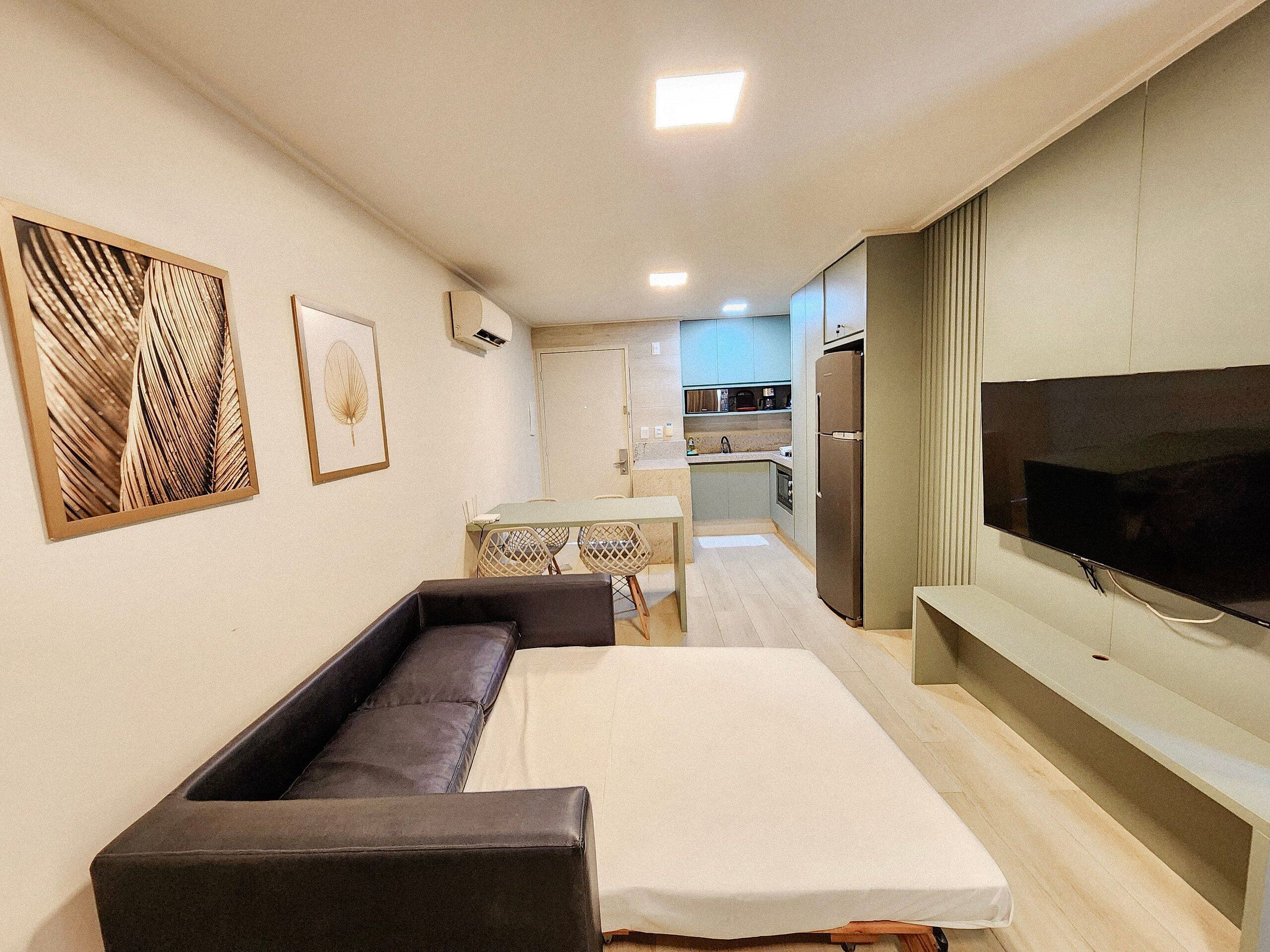 A compact living space with a sofa, a TV mounted on the wall, a dining table with chairs, and a kitchenette in the background.