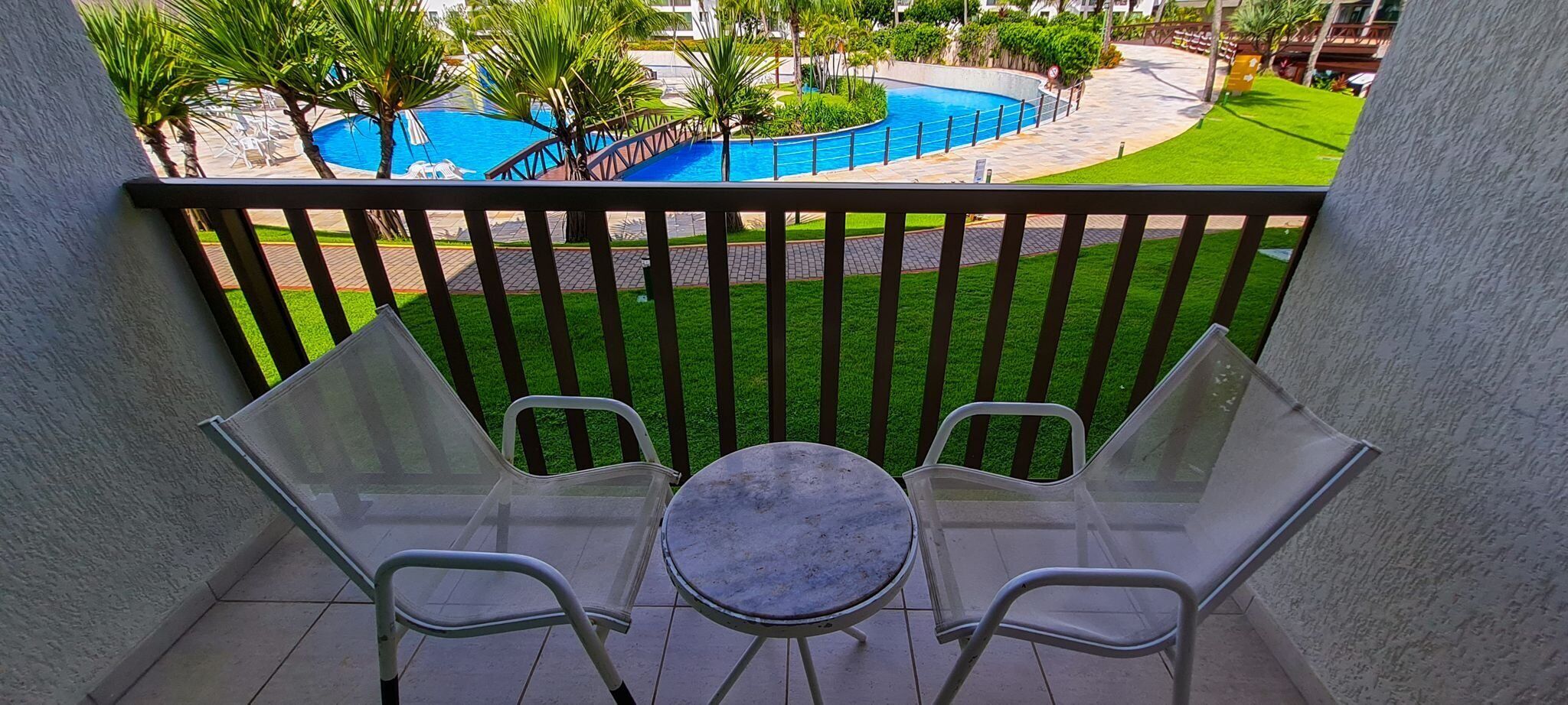 A balcony with two chairs and a small table, overlooking a pool and green lawn.