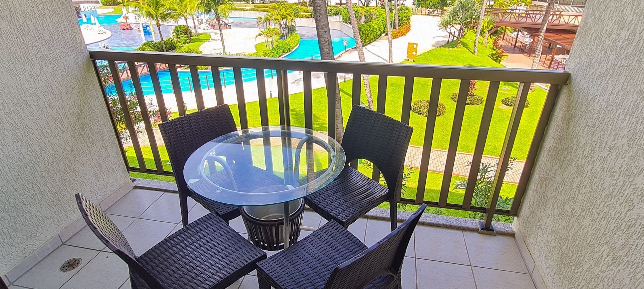 Un balcón con mesa y sillas de vidrio, con vista a una piscina y follaje.