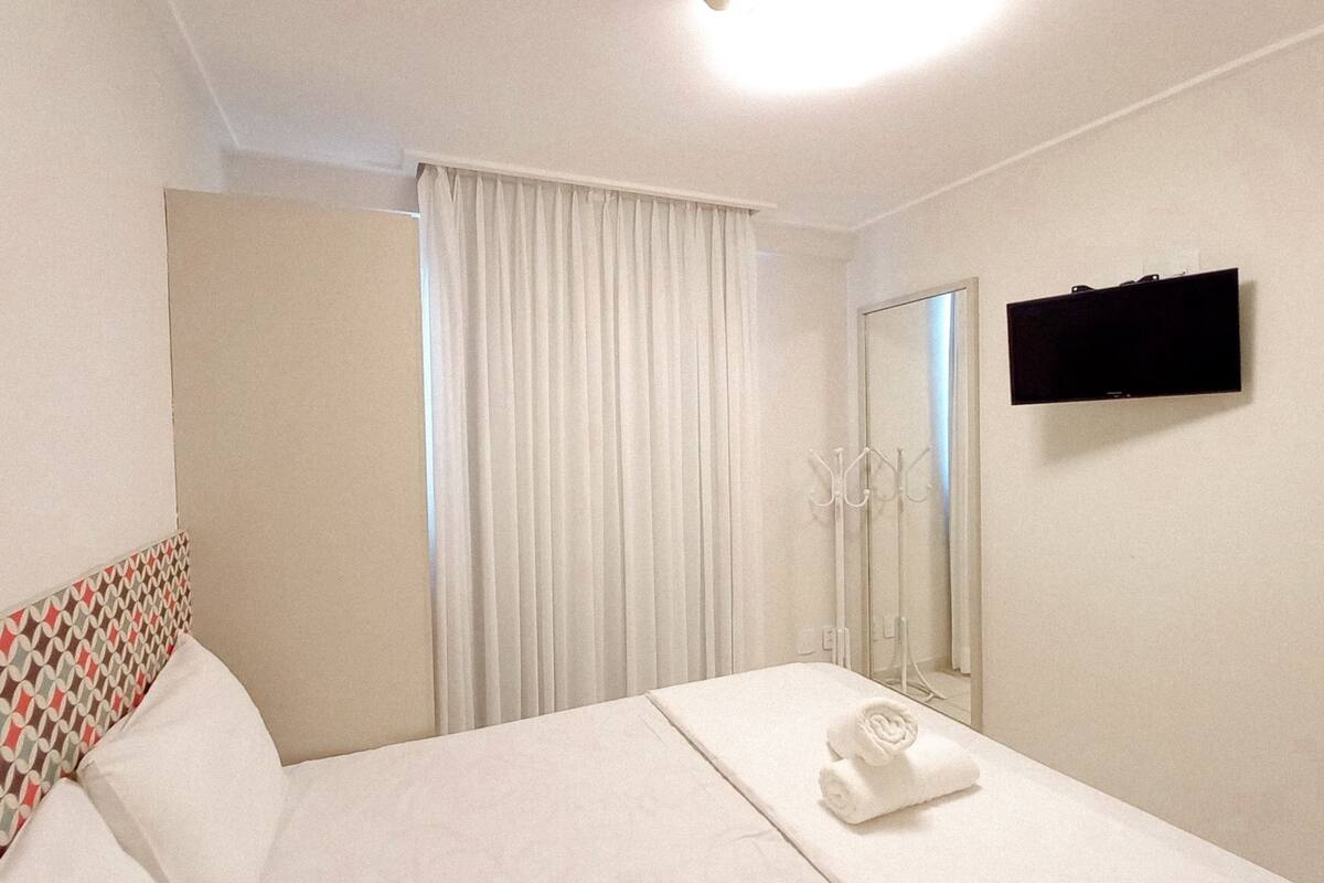 A hotel room with a bed, a TV, a mirror, and a window with curtains.