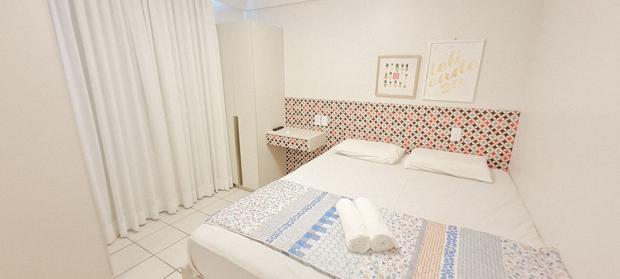 Ein Hotelzimmer mit Bett, Nachttisch, Spiegel, Fenster mit Vorhängen und einer Wand mit gerahmten Bildern.