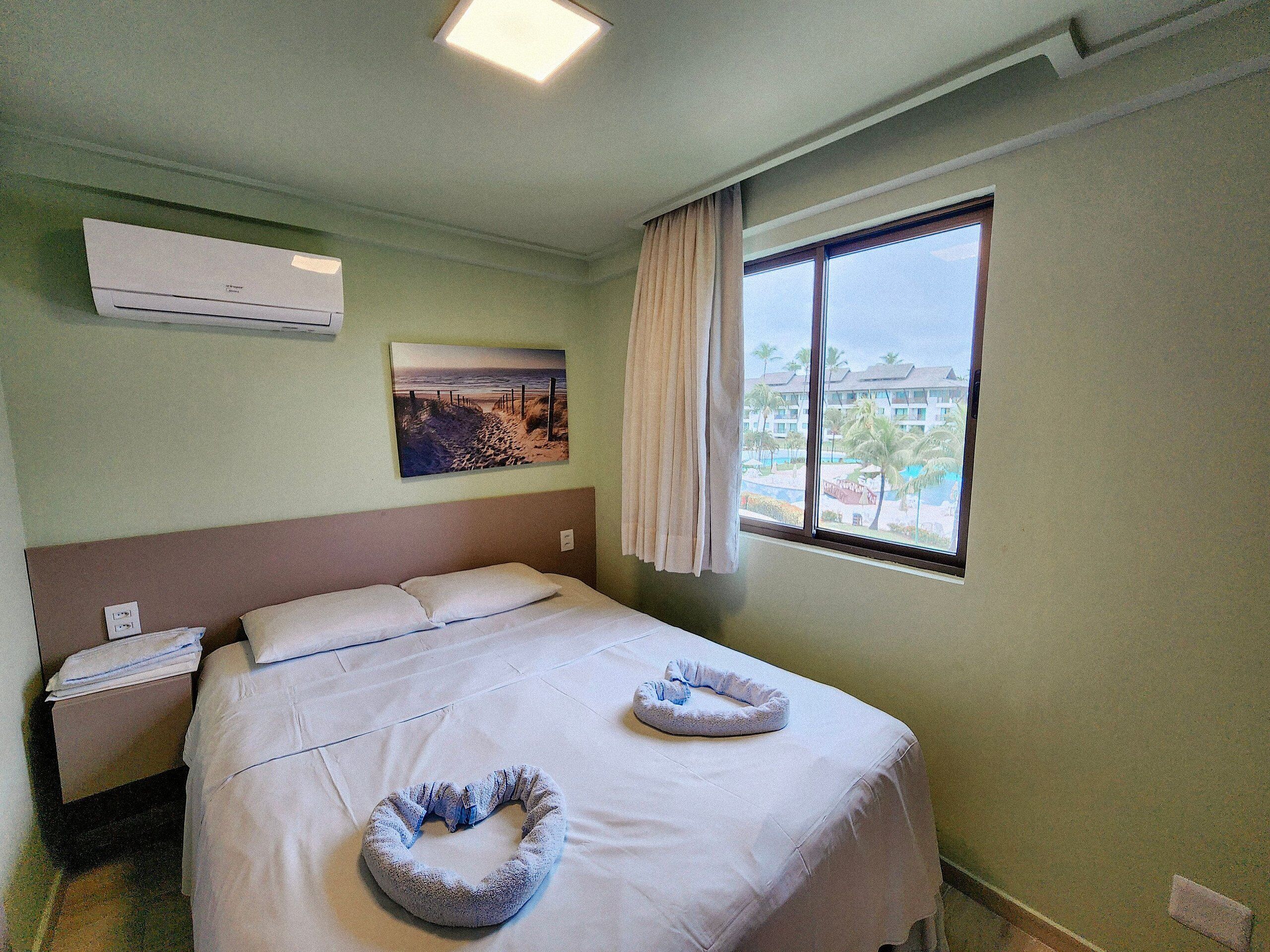 A hotel room with a bed, a window with a view, an air conditioner, and a painting on the wall.
