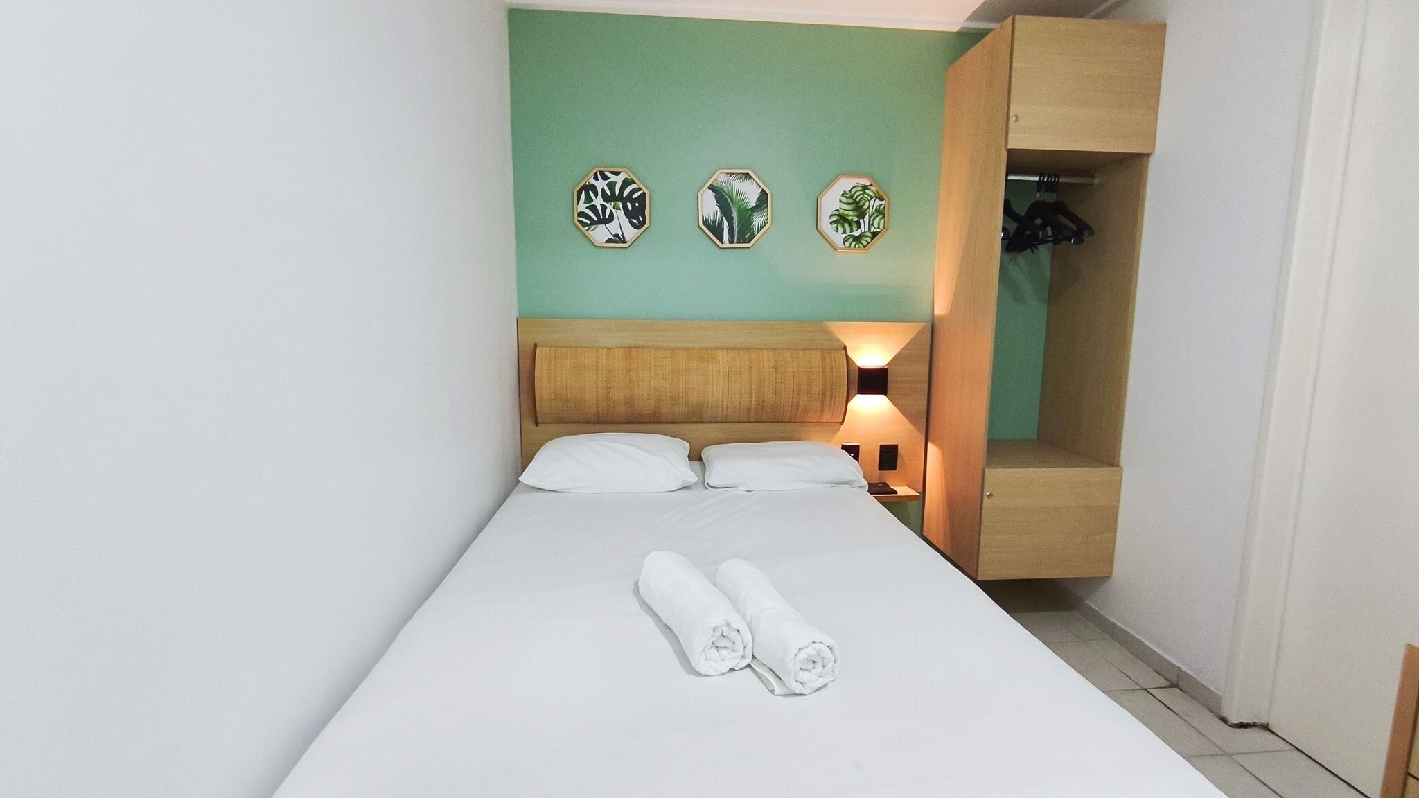 A single bed with white linens, two rolled towels, a wooden headboard, a nightstand with a lamp, and a wooden wardrobe with hanging space.