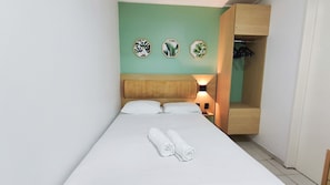 A single bed with white linens, two rolled towels, a wooden headboard, a nightstand with a lamp, and a wooden wardrobe with hanging space. - Flats no Beach Class de Muro Alto (Ipojuca)