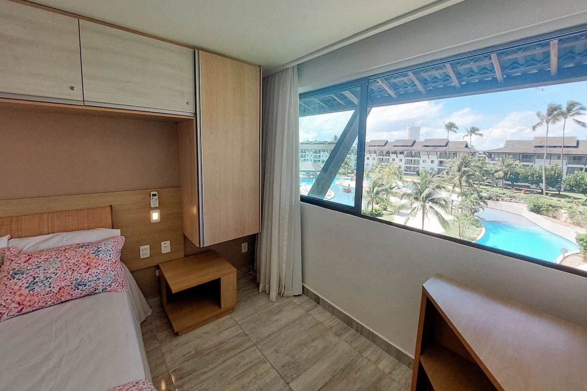 Quarto de hotel com cama, mesa de cabeceira, um banco de madeira, janela com vista para uma piscina e edifícios.