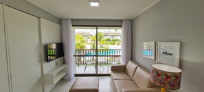 Una sala de estar moderna con sofá, televisor, balcón con vista a la piscina y una mesa con sillas.