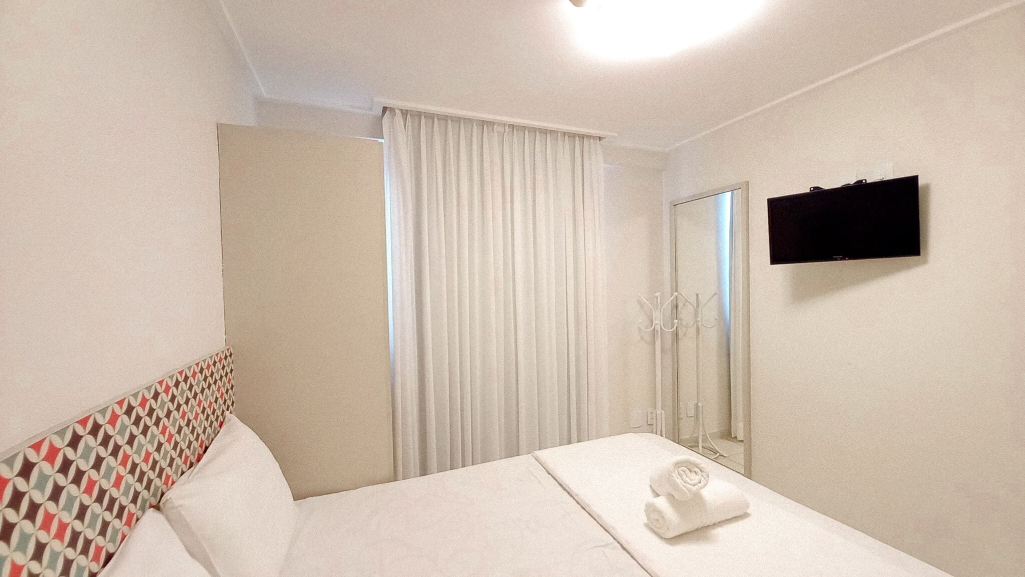 A hotel room with a bed, a TV, a mirror, and a window with curtains.