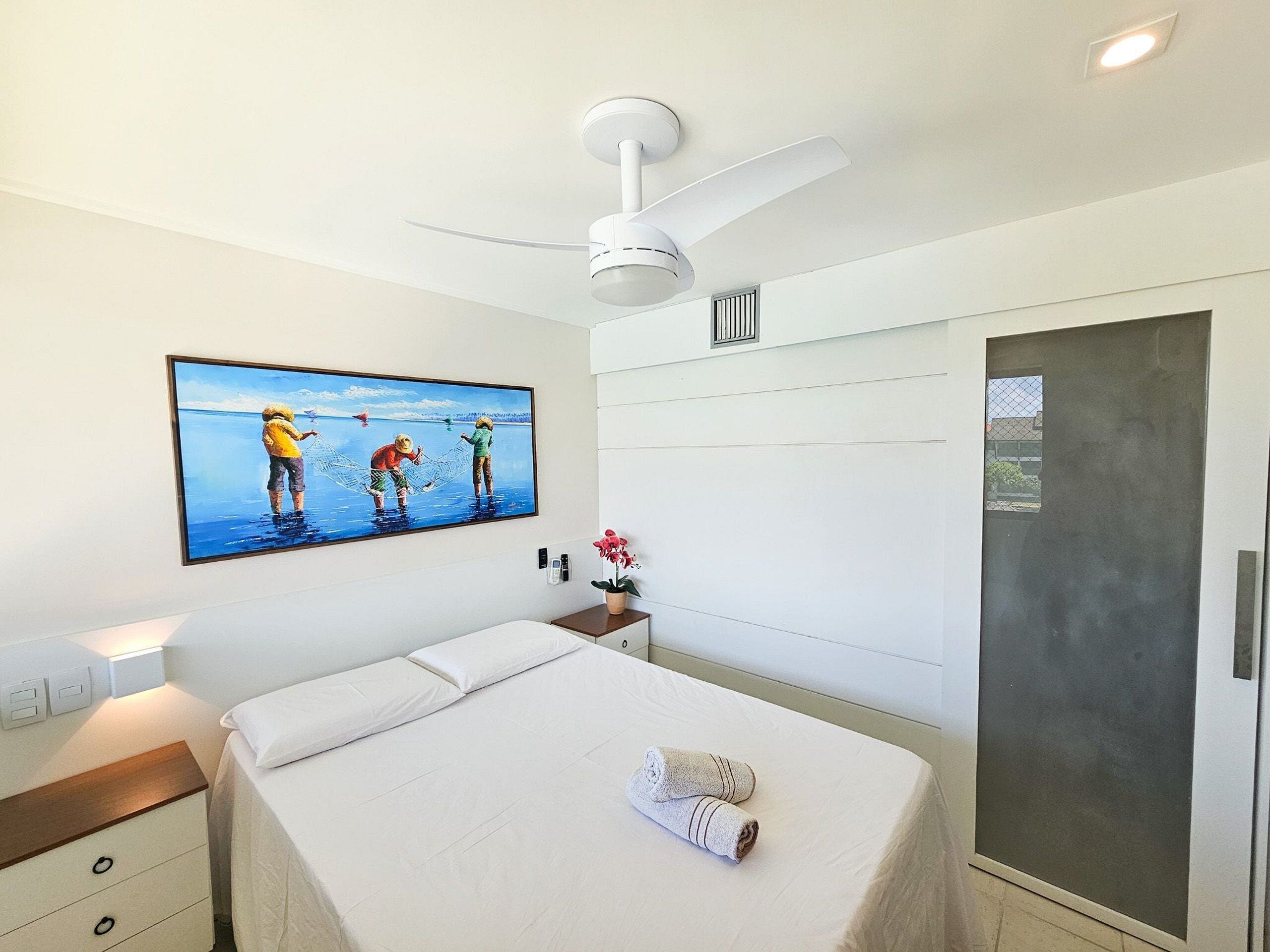 A hotel room with a bed, a nightstand, a painting, a ceiling fan, and a door.
