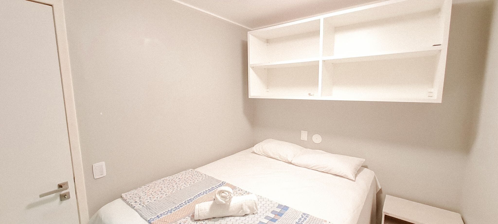 A bedroom with a bed, a bedside table, a wall-mounted shelf, and a door.