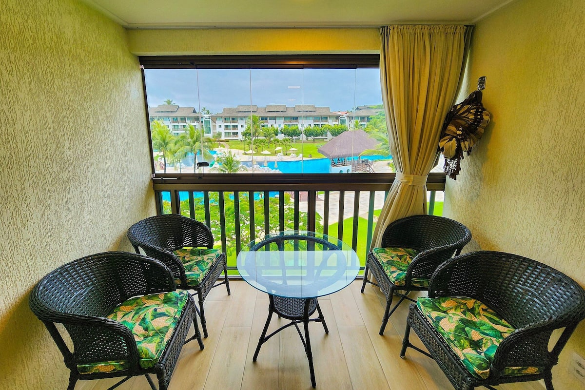 A balcony with wicker chairs and a glass table, overlooking a pool and resort buildings.