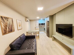 A modern living room with a leather sofa, a flat-screen TV mounted on the wall, and a kitchen area in the background. - Flats no Beach Class de Muro Alto (Ipojuca)
