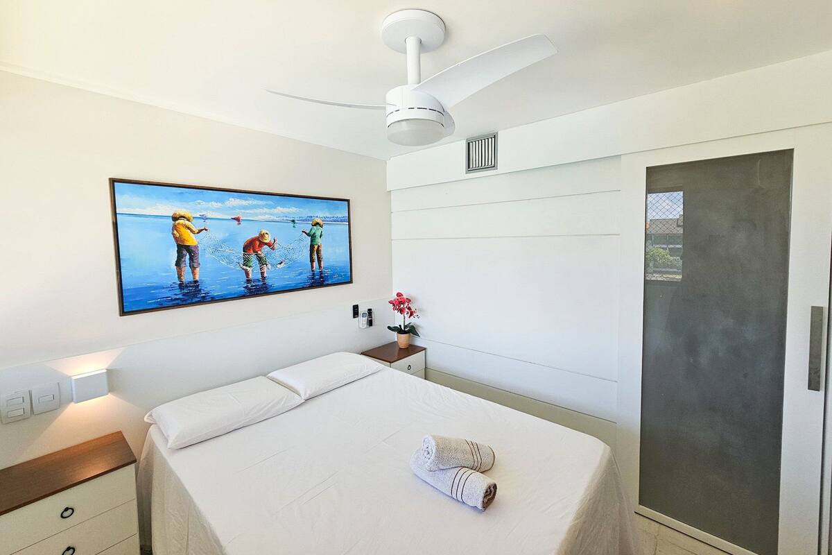 Quarto de hotel com uma cama, mesa de cabeceira, um quadro, ventilador de teto e uma porta.