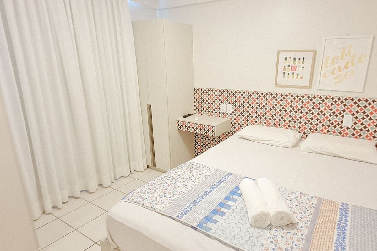 Quarto de hotel com cama, mesa de cabeceira, espelho, janela com cortinas e parede com quadros.