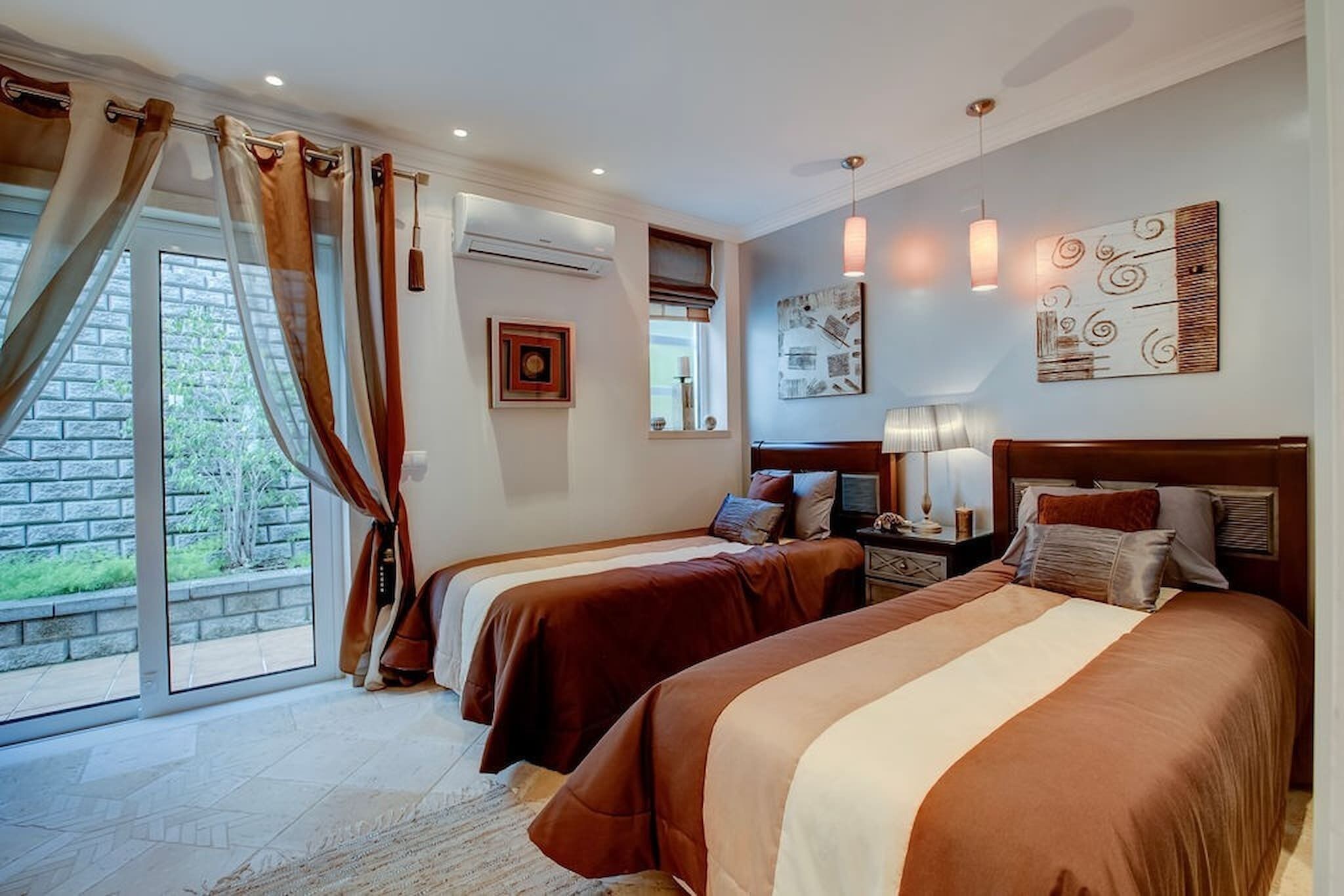 A hotel room with two beds, a large window with curtains, a wall-mounted air conditioner, and a nightstand with a lamp.