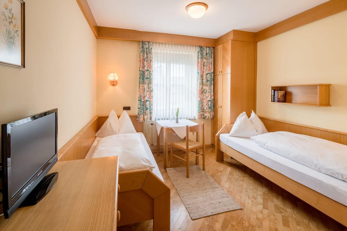 Ein Hotelzimmer mit zwei Einzelbetten, einem Schreibtisch mit Fernseher, einem kleinen Tisch und einem Fenster mit Vorhängen.