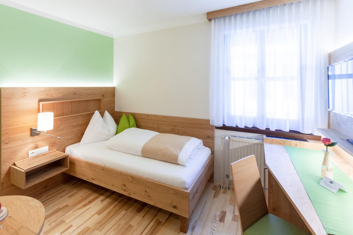 Ein kleines Hotelzimmer mit einem Holzbett, einem Nachttisch mit Lampe, einem Heizkörper und einem Fenster mit durchsichtigen Vorhängen.