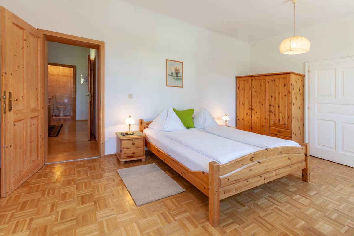Ein hölzernes Bett mit weißen Leinentüchern, ein Nachttisch mit Lampe, ein Holzschrank und eine Tür zu einem Badezimmer.