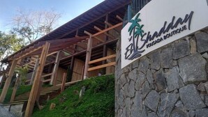 A sign for ElShaddai Boutique with a palm tree logo, wooden structure, and stone wall. - Pousada Recanto El Shadday (Ubatuba)