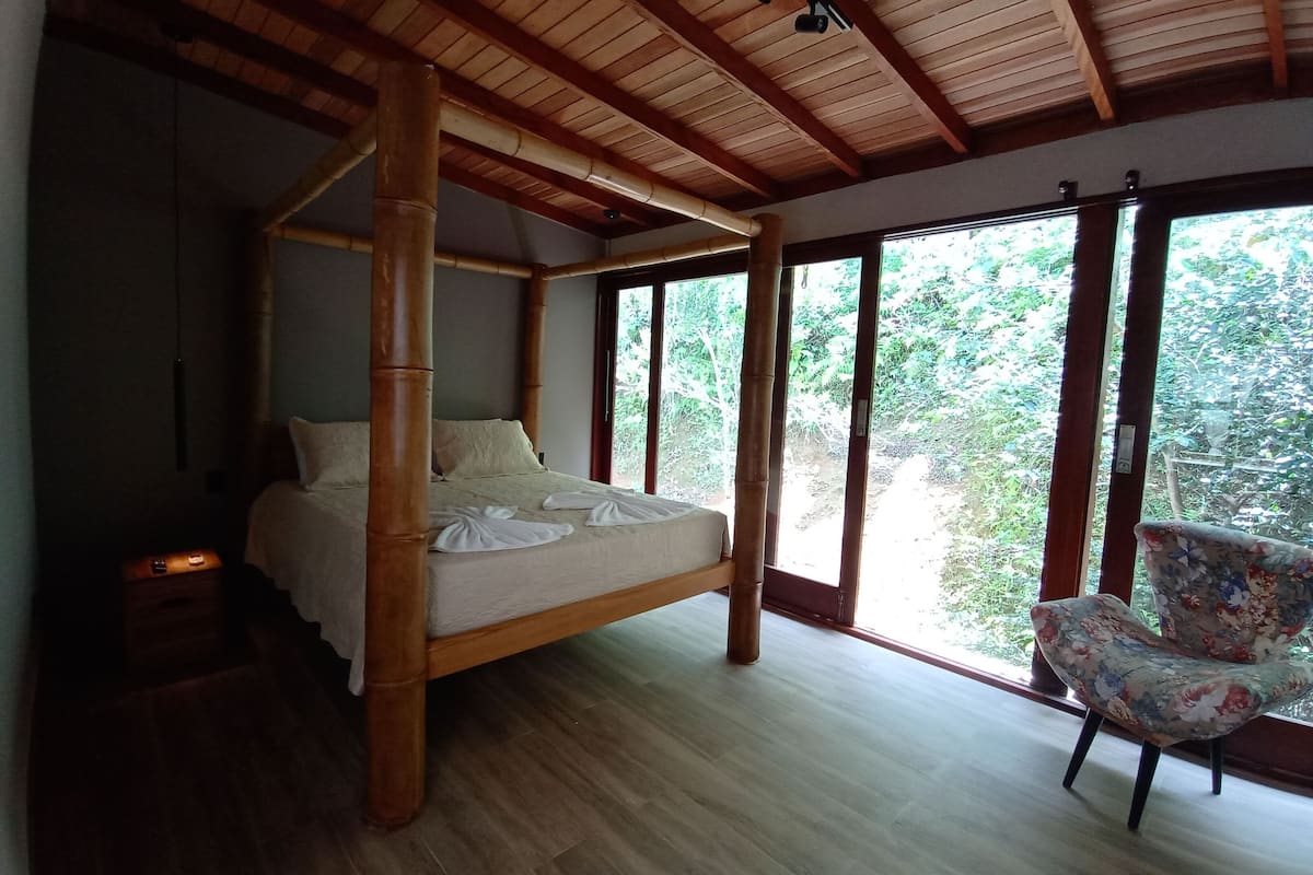 A four-poster bed with a wooden frame and a neutral-colored bedspread. There is a chair with a floral pattern to the right of the bed.