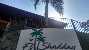 A sign for El Shadday Boutique, featuring a palm tree and a bird, set against a stone wall with a metal fence and a building in the background. - Pousada Recanto El Shadday (Ubatuba)