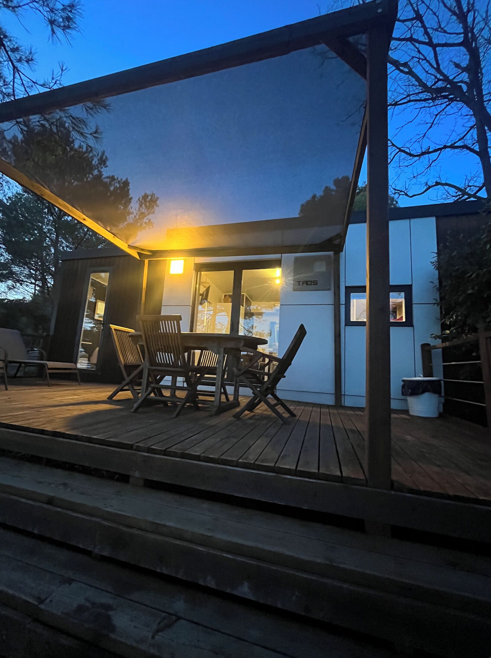 Un área de comedor exterior moderna con una mesa y sillas de madera, un espacio de estar techado y un edificio con un cartel que dice "TAOS".
