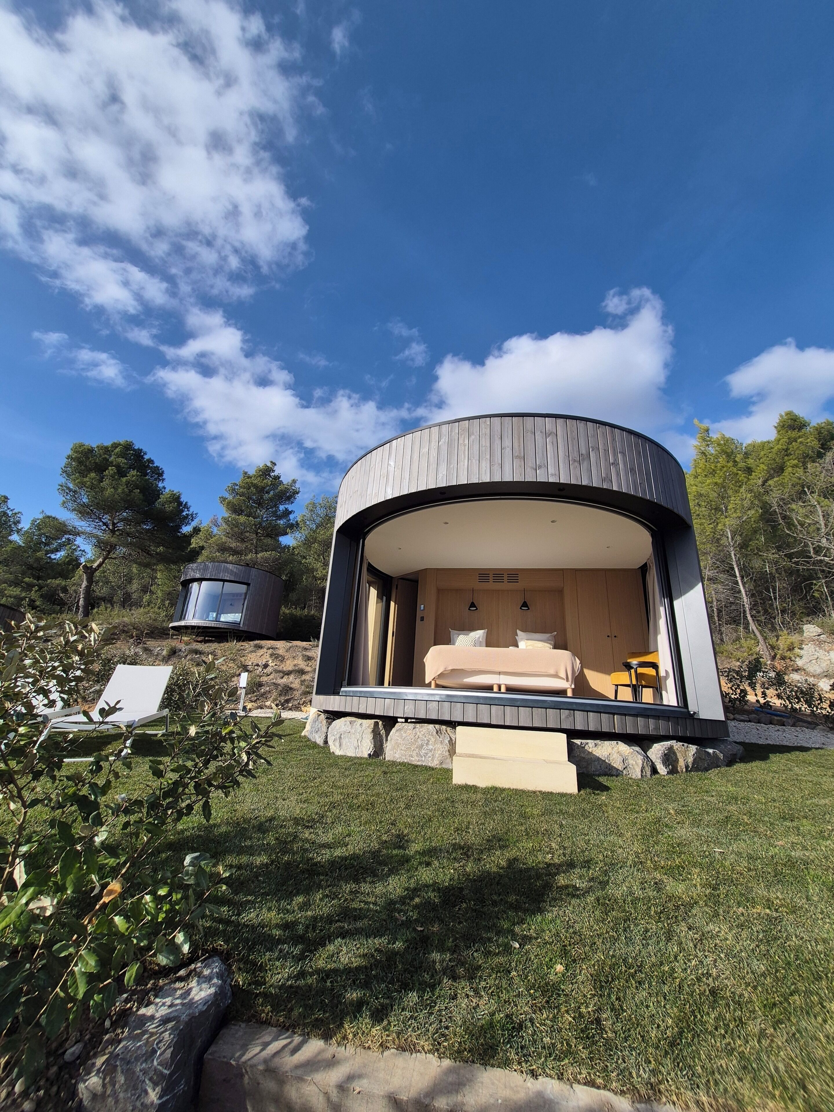 A modern, circular building with a large glass window, a bed, and a chair visible inside.