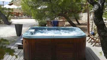 Un jacuzzi en una terraza de madera con una pared de piedra y mobiliario exterior al fondo.