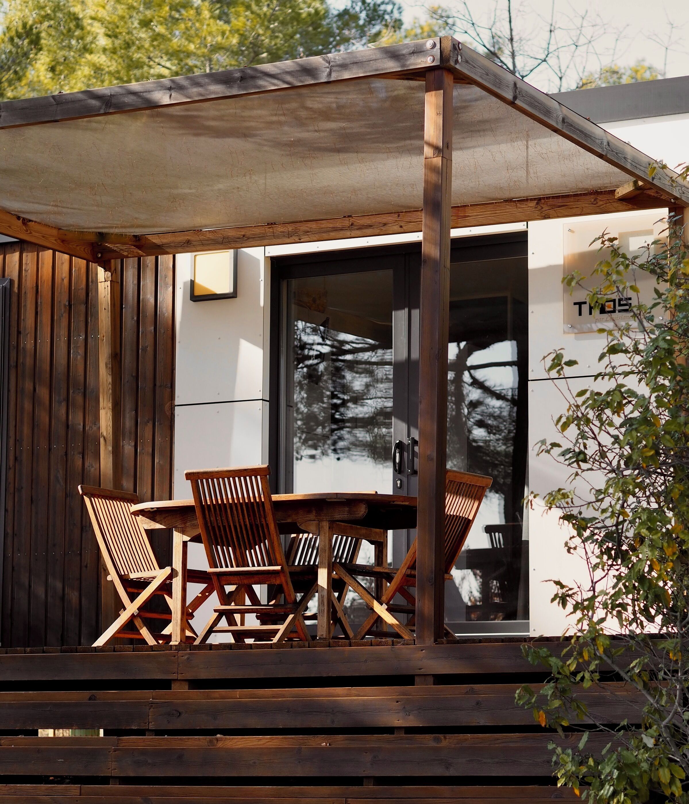 Une terrasse en bois avec une table et des chaises, un espace couvert et un bâtiment doté d’une porte en verre et d’une enseigne indiquant « TYOS ».