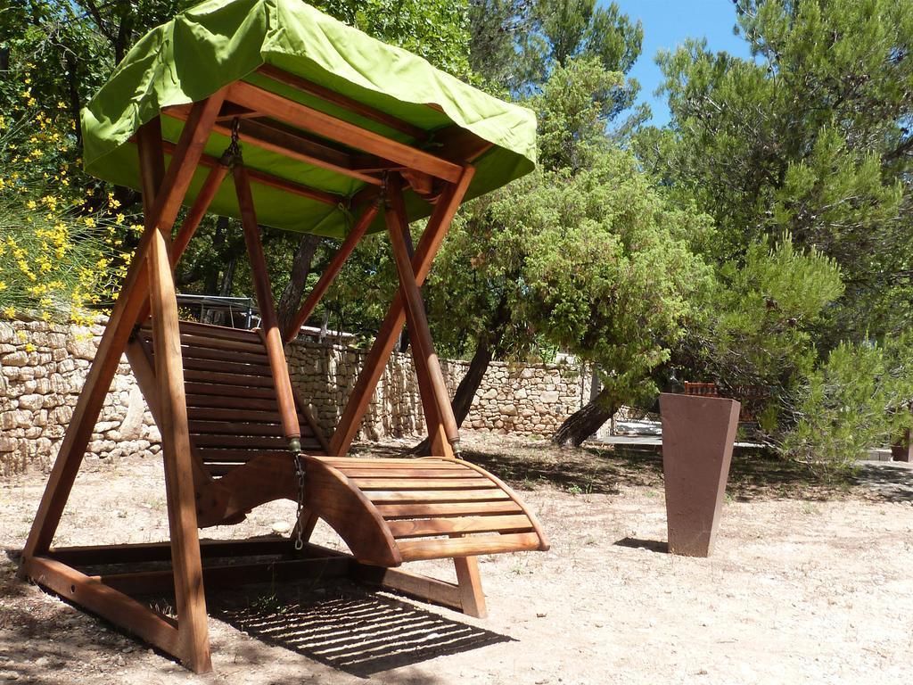 A wooden swing chair with a green canopy, set against a backdrop of trees and a stone wall.