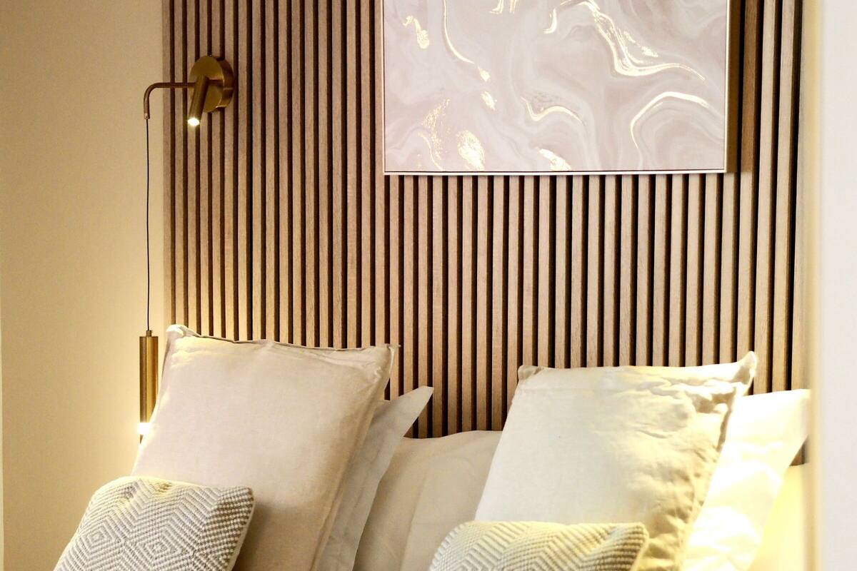 Une chambre moderne avec un lit, des oreillers, une lampe fixée au mur et une œuvre d’art encadrée.