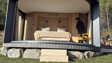 Eine moderne, runde Holzhütte mit einem Bett und einem Stuhl im Inneren, eine Person steht an der Tür, und ein klarer Himmel ist im Hintergrund zu sehen.
