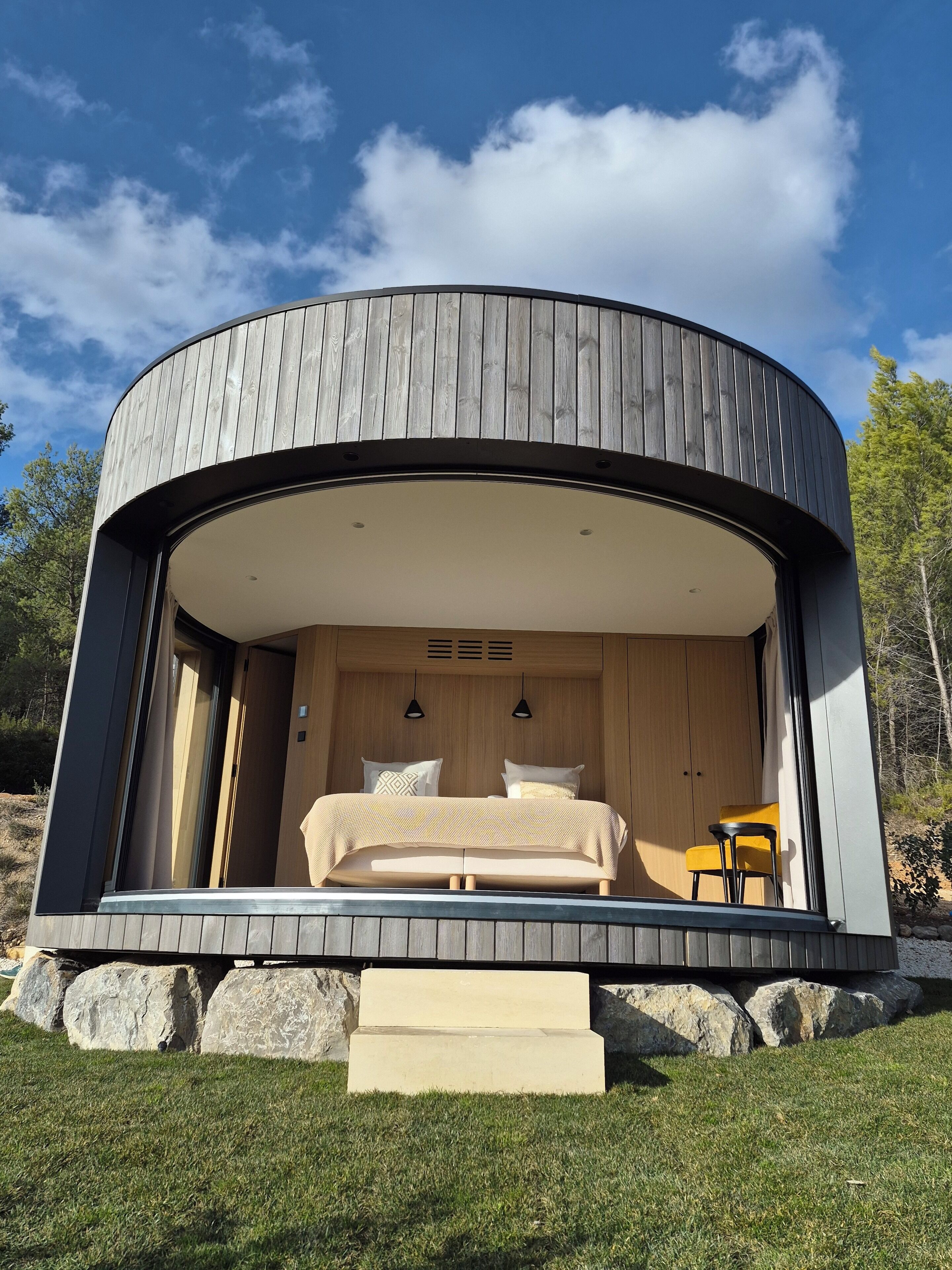 A modern, circular wooden cabin with a bed, two bedside tables, and a small chair.