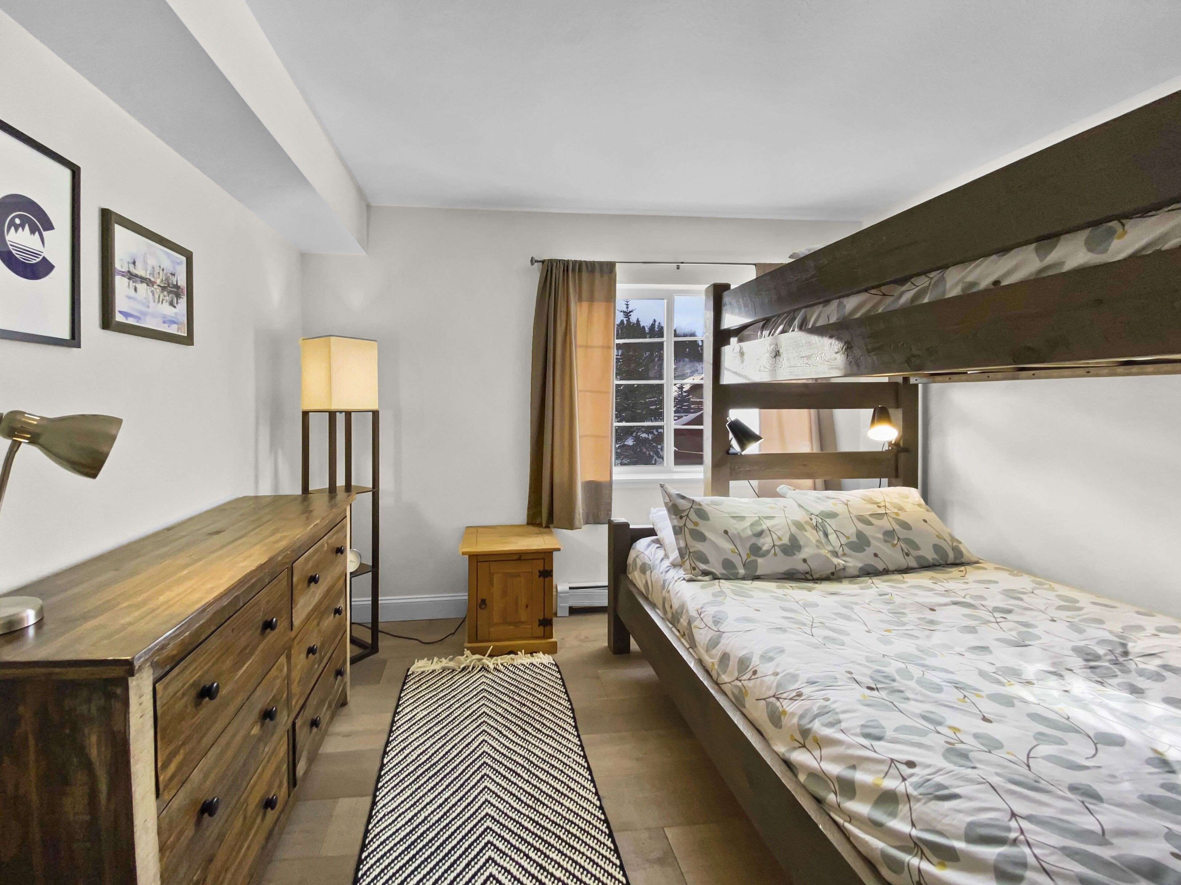Une chambre avec un lit superposé, une commode en bois, un bureau, une lampe et une fenêtre avec des rideaux.