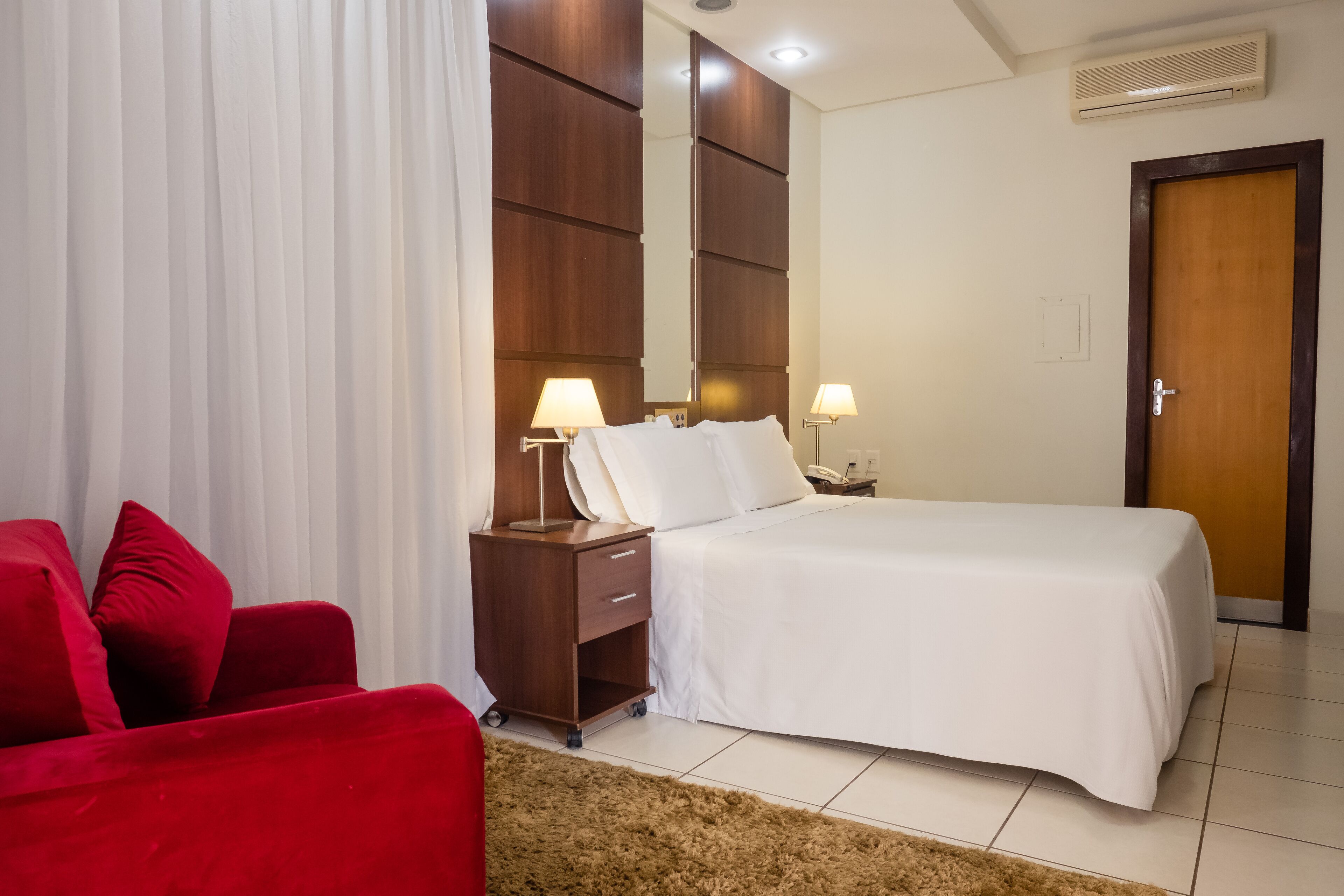 A hotel room with a bed, a red sofa, a wooden nightstand with lamps, and a door.