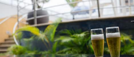 Zwei Biergläser auf einer spiegelnden Oberfläche, im Hintergrund verschwommen Grün und ein Gebäude.