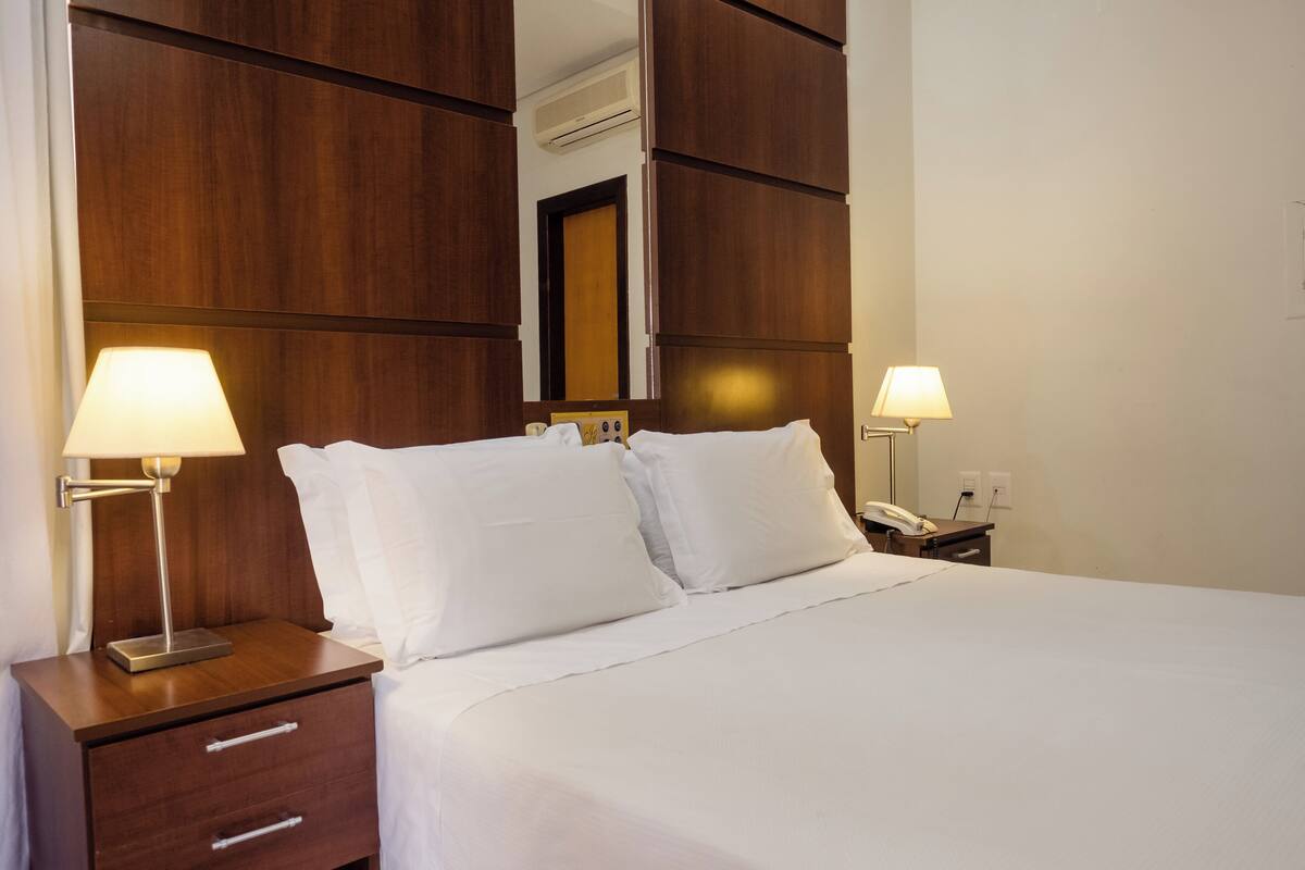 A hotel room with a wooden headboard, two bedside lamps, a nightstand with drawers, and a large bed with white linens.