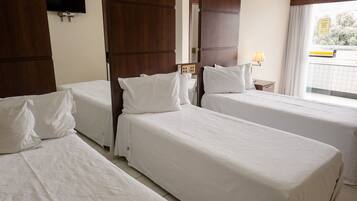 A hotel room with two single beds, a TV mounted on the wall, a window with a view, and a bedside table with a lamp.