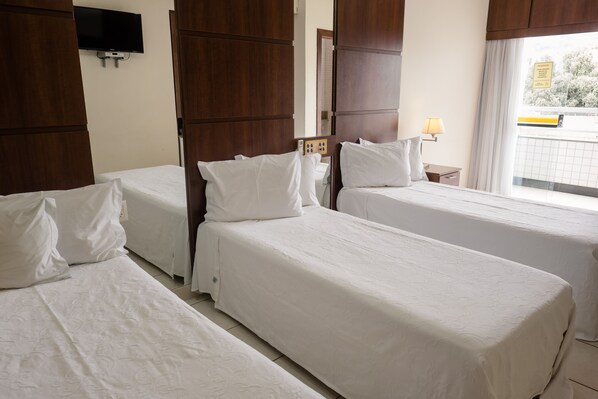 A hotel room with two single beds, a TV mounted on the wall, a window with a view, and a bedside table with a lamp.