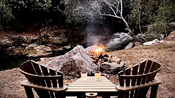 A wooden table with two Adirondack chairs in a forested area with a campfire.