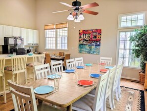 Un coin repas avec une table en bois dressée pour six personnes, des chaises blanches, un ventilateur de plafond et une fenêtre donnant sur un beau paysage.