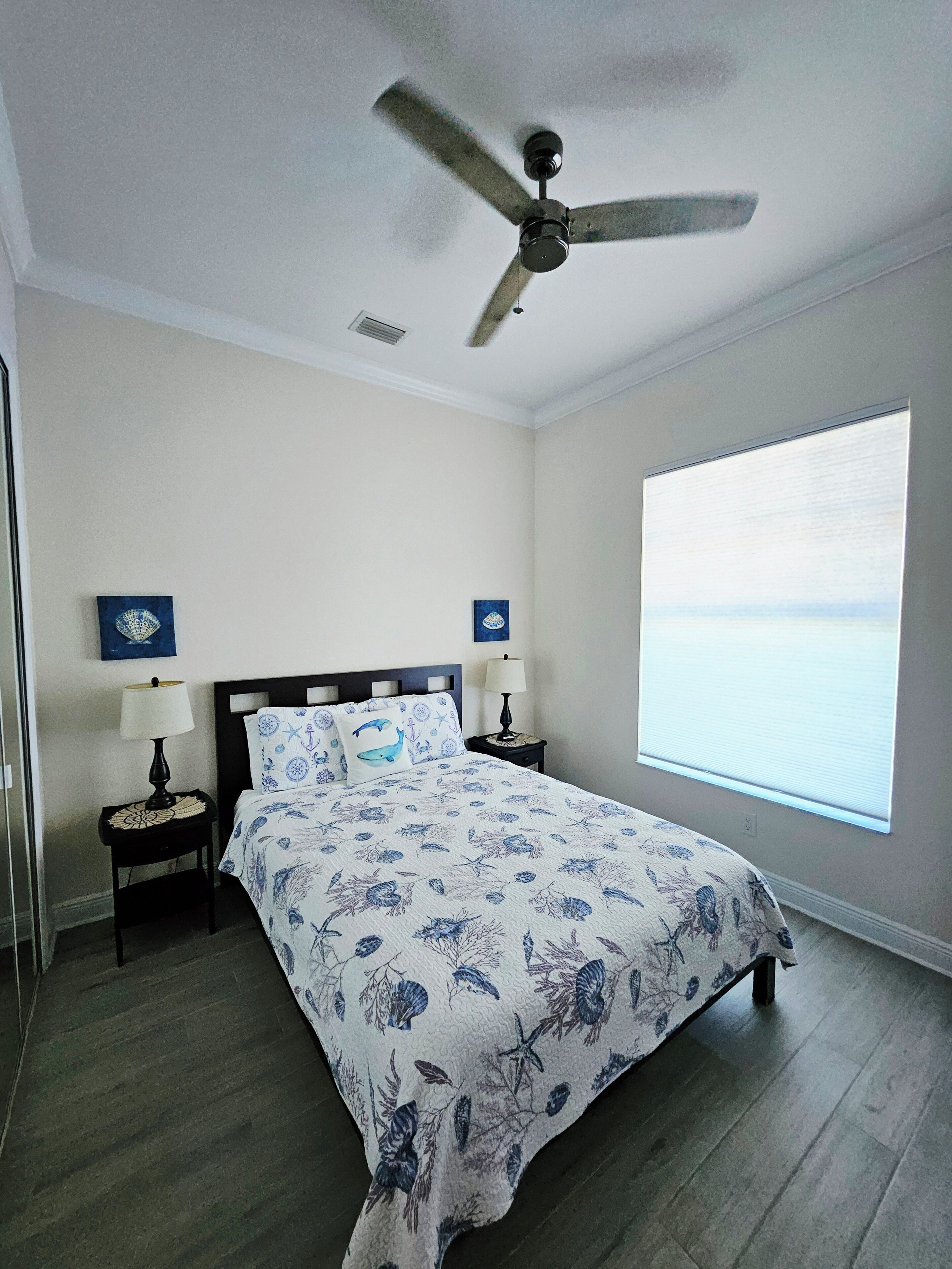 A bedroom with a bed, a ceiling fan, a nightstand with a lamp, a window with a view, and a wall with a painting.