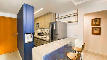 Uma cozinha compacta com geladeira embutida, máquina de café e micro-ondas. Há uma pequena ilha com banquetas.