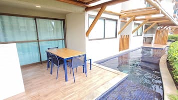 A poolside area with a wooden deck, a table with chairs, and a glass-enclosed room.