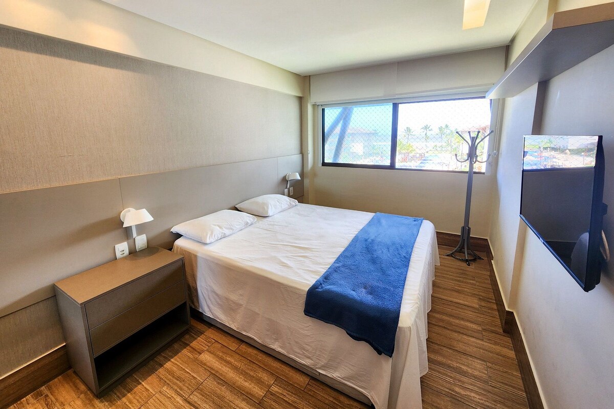 A modern hotel room with a bed, bedside table, lamp, and a TV mounted on the wall.