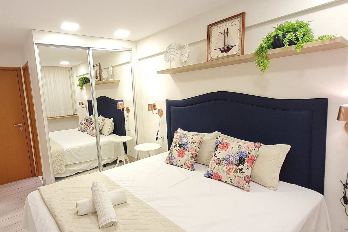Un dormitorio con cama, mesita de noche, espejo y una ventana pequeña.