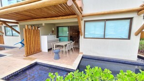 A pool area with a tiled edge, a covered patio with a table and chairs, and a view of the interior through a glass door. - Flats no La Fleur Polinésia Residence (Ipojuca)