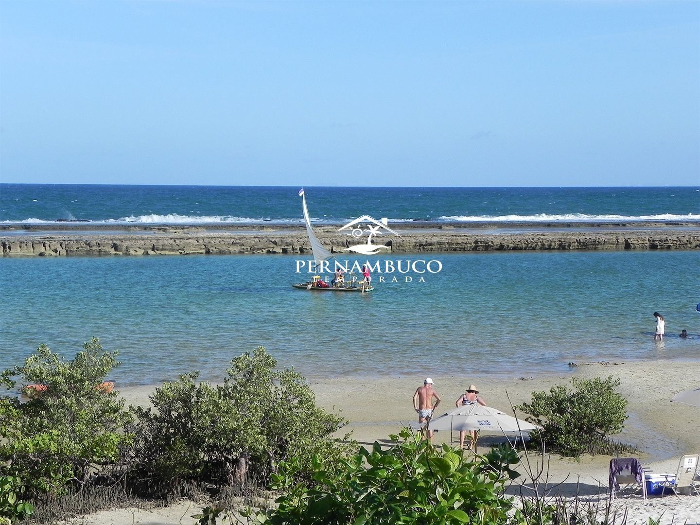 Una playa con costa rocosa, un velero y gente en la arena.