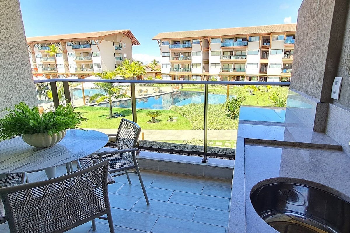 Un balcón con mesa, sillas, un lavabo y vistas a una piscina y edificios de apartamentos.